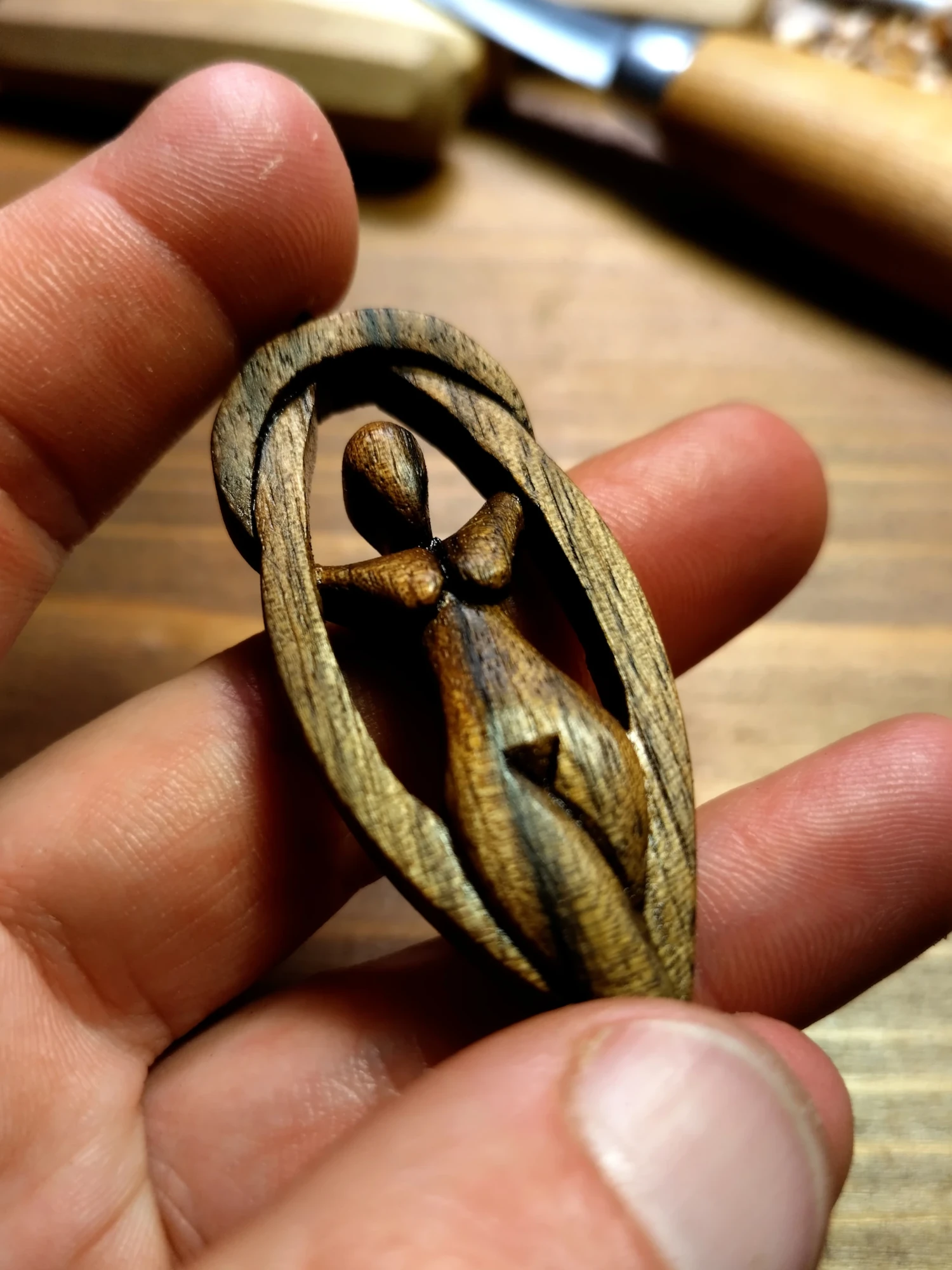 Frisch geölter Göttin Gaia Holzanhänger, handgeschnitzt aus dunklem Nussbaumholz in einer Hand. Fotografiert vor einer Werkbank mit Holzspänen und Schnitzmessern im Hintergrund.