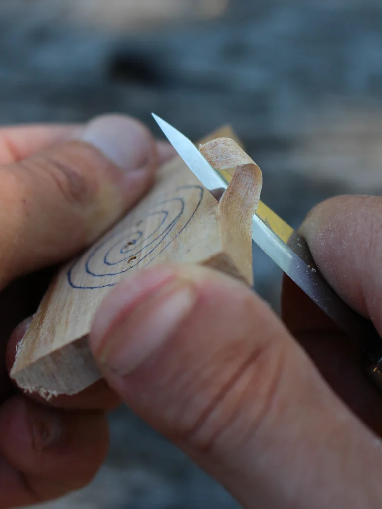 Nahaufnahme einer Hand, die mit einem kleinen Schnitzmesser eine feine Holzspäne aus einem Stück Restholz löst; auf dem Holz sind vorgezeichnete Linien erkennbar. Das Bild zeigt den Beginn handgemachter Naturschmuck-Herstellung – ruhige, achtsame Handarbeit ohne Maschinen.