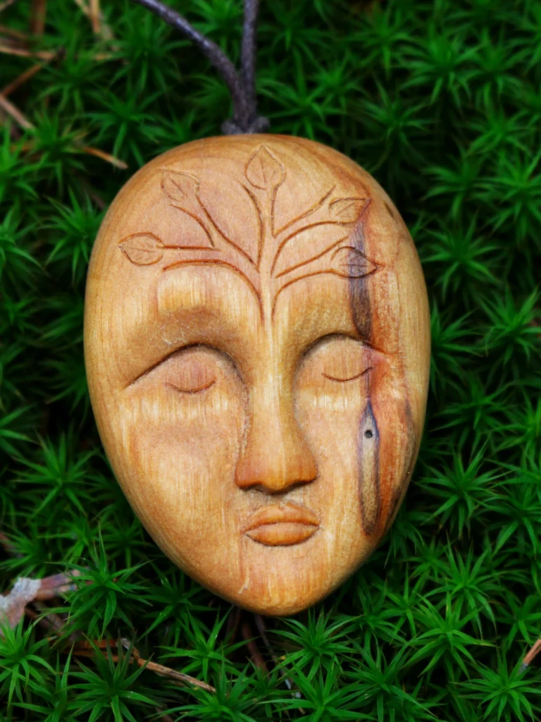 Handgeschnitzter Talisman aus gesammeltem Birkenholz auf natürlicher Moosunterlage im Wald. Gesicht mit geschlossenen Augen auf der Stirn verziert mit Blattranken und einer markanten Zeichnung unter dem rechten Auge, die an Träne erinnert unter dem Auge.